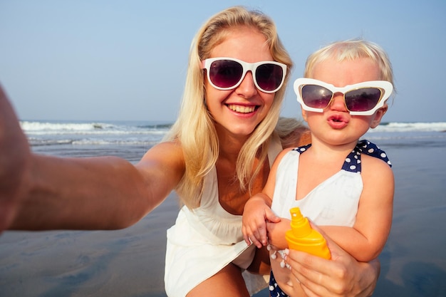 Fairhaired mother in stylish glasseswhite dress orange spray bottle SPF sunscreen with newborn baby one year old lips duck air kiss daughter taking portrait on the camera smartphone summer beach