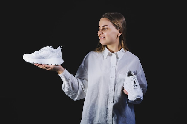 A fairhaired woman in a white shirt on a black background holds white sneakers made of genuine leather in her hands