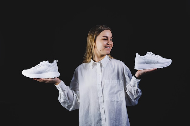 A fairhaired woman in a white shirt on a black background holds white sneakers made of genuine leather in her hands