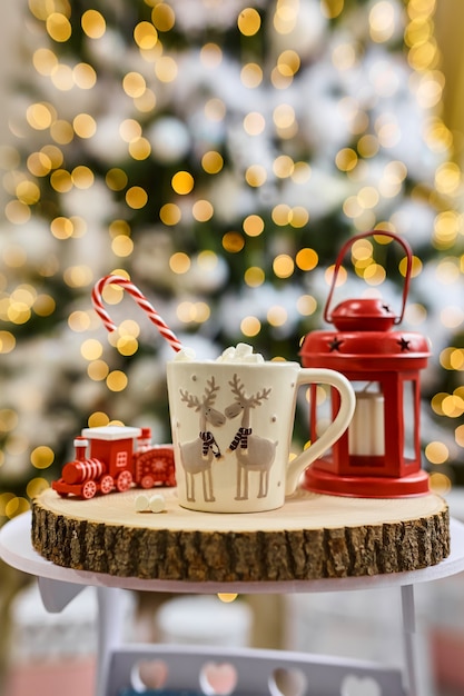 Fairy lights background, hot cocoa with marshmallows, white cup with deer on a wooden cut and red lantern and children's train at home during Christmas and New Year holidays. Soft focus bokeh.