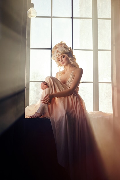 光の中で長いブロンドの髪を持つ妖精は、大きな窓、アートファッションの女の子の背景にあります。長いベージュのドレスを着て窓に日光の下で座っている美しいブロンドの女性