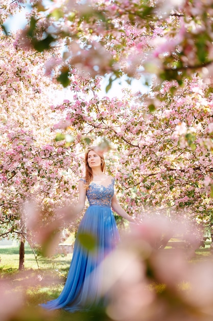 Ragazza da favola con i capelli rossi in un abito da sera blu passeggiate nel giardino di fiori di ciliegio rosa in una giornata di sole sfocato concetto anallergico in primo piano