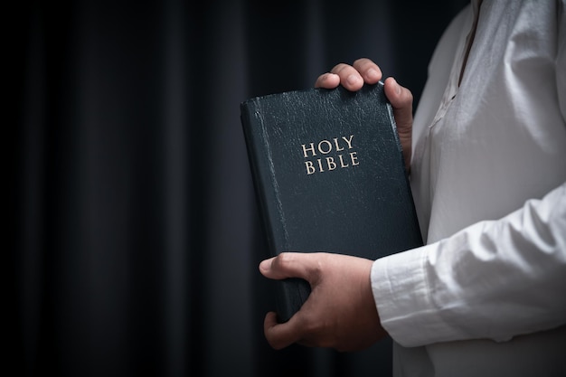 Faith with holy bible concept hands of a female prayer worship\
god with holy bible on black background in church christian woman\
who believe in jesus read and study the grace of the holy\
scriptures