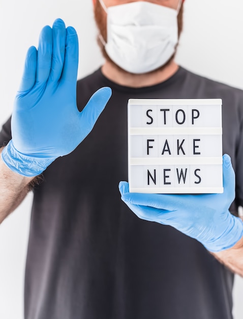 Fake news infodemics during Covid-19 pandemic concept. Man wearing protective mask and medical gloves on hands holding lightbox with text Stop fake news. People want to know truth about coronavirus