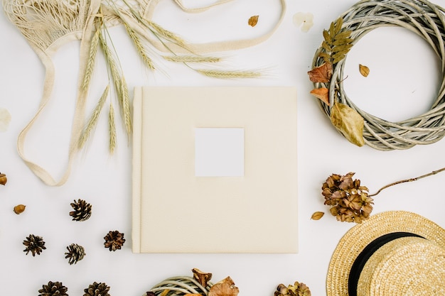 Fall autumn composition with wedding or family photo album, wreath frame, string bad, rye ears, cones, dry leaves, straw hat on white surface