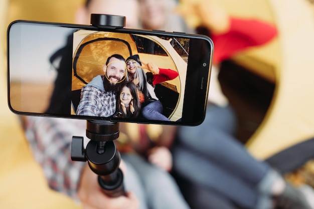 秋のキャンプテントに座っている両親と娘と一緒に画面のクローズアップスマートフォンのカメラを使用して自分撮りをする若い家族