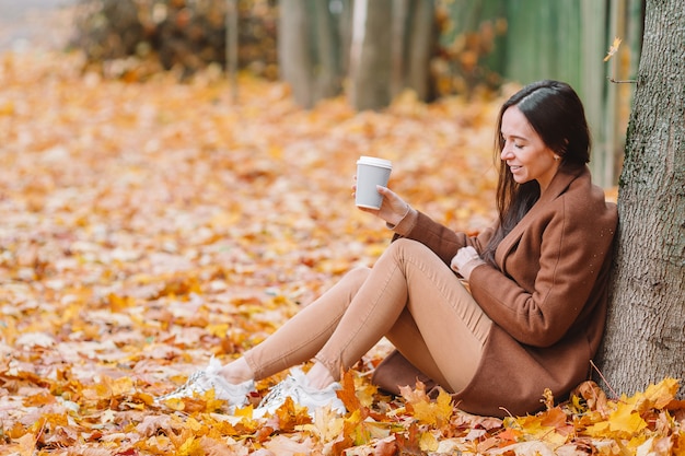 Fall concept - beautiful woman drinking coffee in autumn park under fall foliage