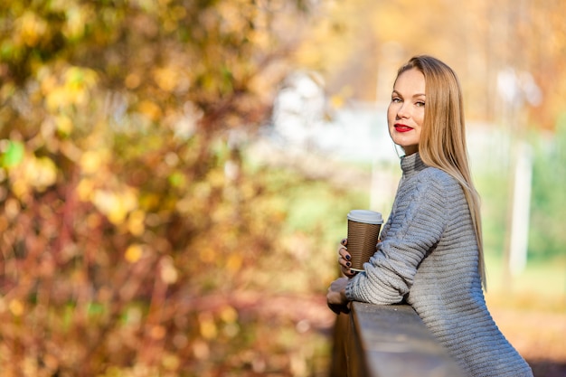 Fall concept - beautiful woman drinking coffee in autumn park under fall foliage