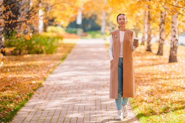 Fall concept - beautiful woman drinking coffee in autumn park under fall foliage