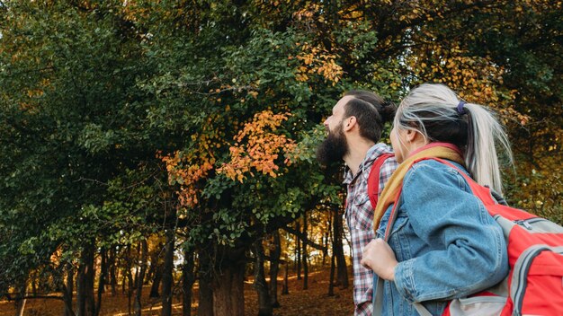 Fall journey Side view of couple traveling with backpacks in autumn nature park Green trees and yellow foliage background