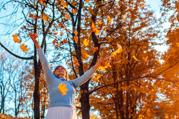 Fall season. Woman throwing leaves in autumn forest. Senior woman having fun outdoors