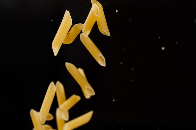 Falling penne pasta. Flying yellow raw macaroni over black background. Shallow dof.