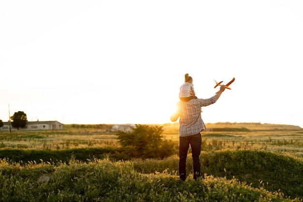 Familie alleenstaande vader met zijn dochter bij zonsondergang in een veld op een zomeravond die een speelgoedvliegtuig in de lucht lanceert babymeisje zittend op papa's schouders kijkt naar het skychildhood-concept