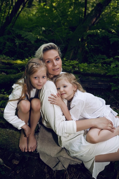 Foto familie blonde moeder met twee kinderen in een witte jurk ligt op boomstam in het bos blootsvoets