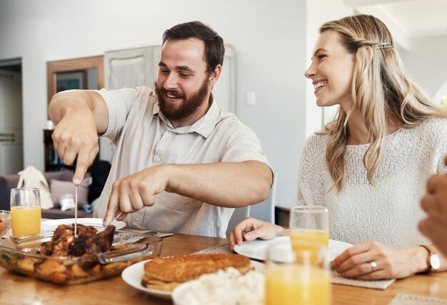 Foto familie eten en man die kip snijdt tijdens een etentje met lachende vrouw die samen eet en drinkt in de eetkamer gelukkig zorg en deel tijd met koppel en vrienden in huis in canada