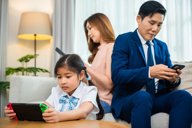 Familie geeft niets om elkaar. Aziatische ouders negeren hun kind en kijken thuis naar hun mobiele telefoon, vader en moeder lezen sociale media maar dochter speelt videogame op sofa woonkamer