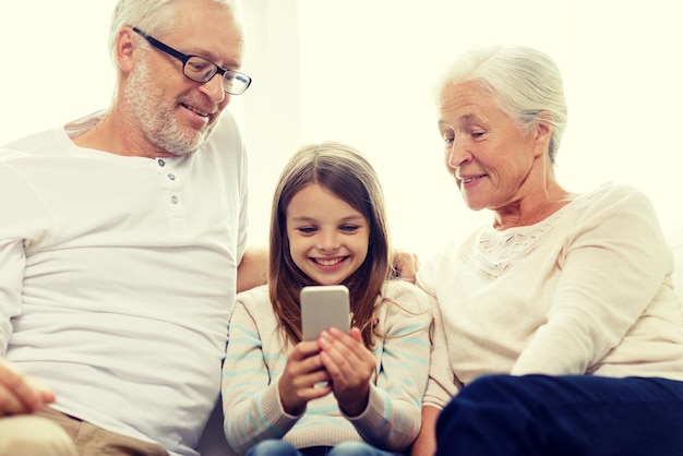 familie, generatie, technologie en mensenconcept - glimlachende grootvader, kleindochter en grootmoeder met smartphone die thuis op de bank zit