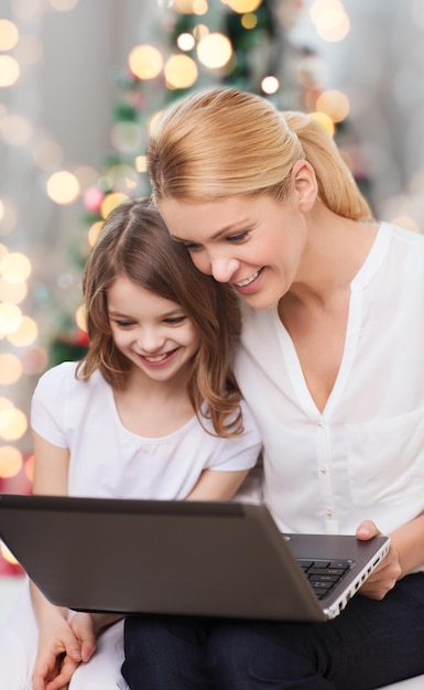 familie, jeugd, vakanties, technologie en mensen - glimlachende moeder en klein meisje met laptopcomputer over de achtergrond van kerstboomverlichting