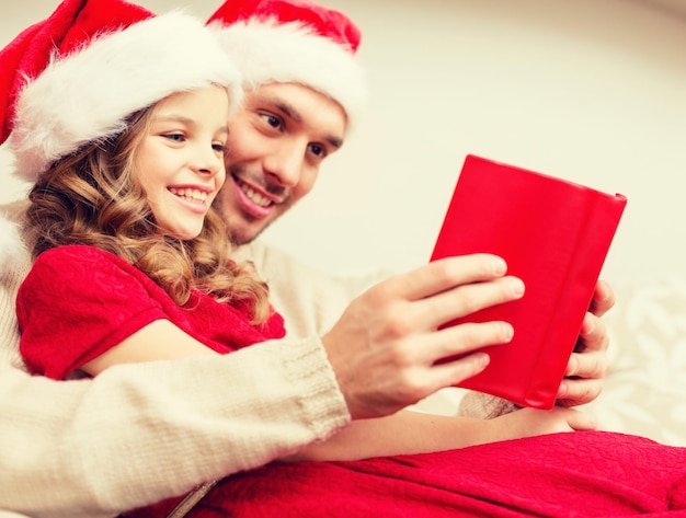 familie, kerstmis, x-mas, geluk en mensenconcept - lachende vader en dochter in kerstman helper hoeden leesboek
