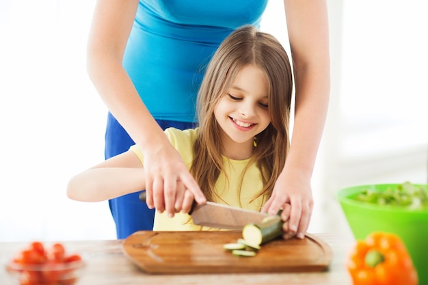 familie, kind, koken en thuisconcept - lachend meisje met moeder die komkommer hakt in de keuken