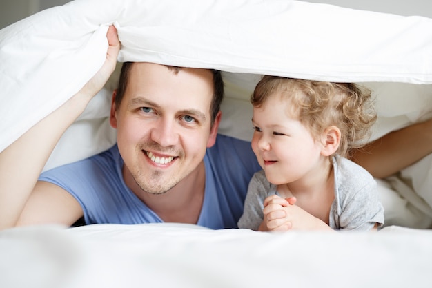 Familie, liefde, geluk en vaderschap concept - portret van gelukkige vader en schattige kleine meisjesdochter liggend in bed onder deken