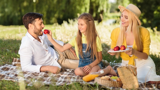 Foto familie met picknick in het park na quarantaine, dochter voedt vader met appel en geniet samen van zomerdag. panorama