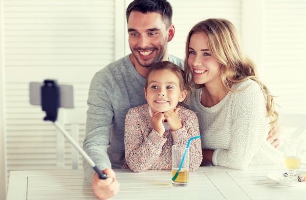 familie, ouderschap, technologie en mensen concept - gelukkige moeder, vader en klein meisje die dineren en foto's maken met een smartphone selfie stick in een restaurant