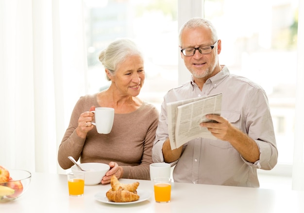 Familie, technologie, eten, drinken en mensen concept - gelukkig senior paar ontbijten en krant lezen thuis