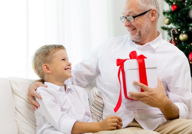 familie, vakantie, generatie, kerst en mensen concept - lachende grootvader en kleinzoon met geschenkdoos zittend op de bank thuis