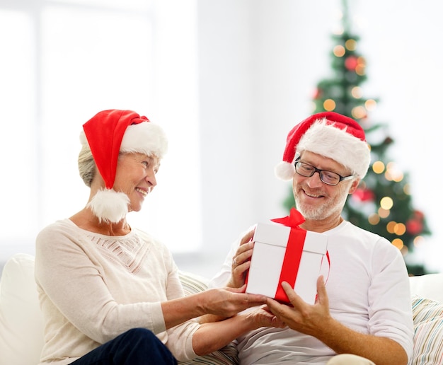 familie, vakantie, leeftijd en mensen concept - gelukkig senior paar in santa helper hoeden met geschenkdoos over woonkamer en kerstboom achtergrond
