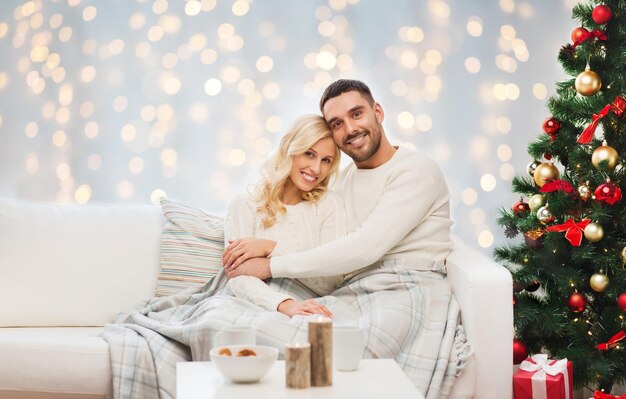 familie, vakantie, liefde en mensen concept - gelukkige paar bedekt met geruite zittend op de bank over kerstboom en lichten achtergrond