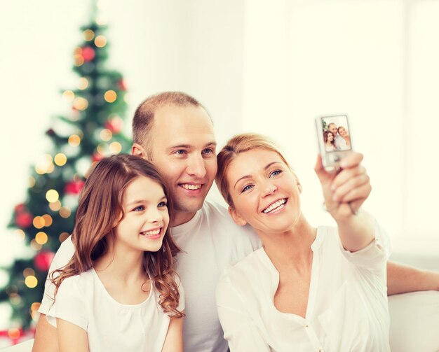 familie, vakantie, technologie en mensen - glimlachende moeder, vader en klein meisje maken selfie met camera over woonkamer en kerstboomachtergrond