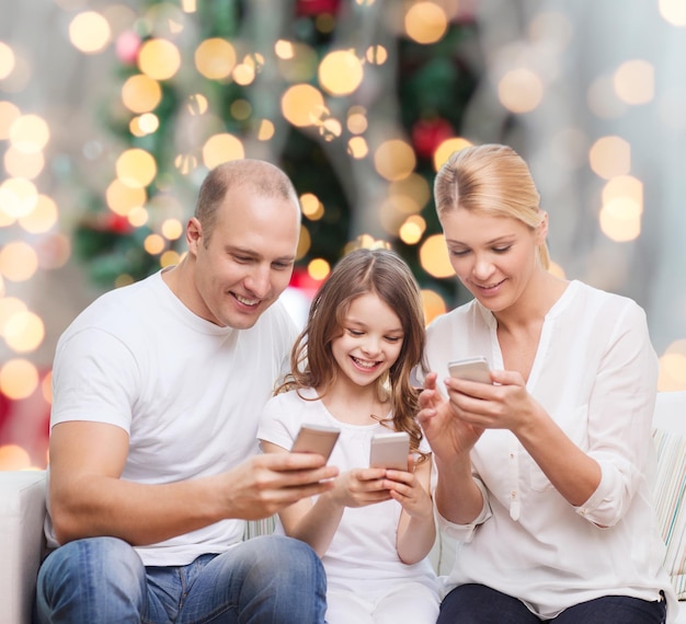 familie, vakantie, technologie en mensenconcept - glimlachende moeder, vader en klein meisje met smartphones over woonkamer en kerstboomachtergrond