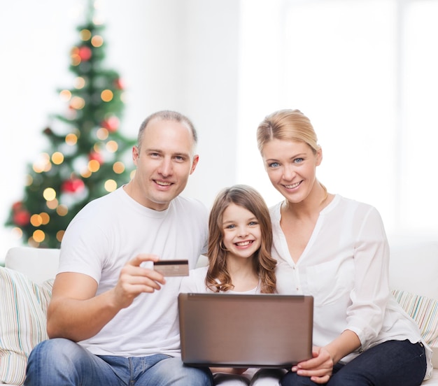 familie, vakantie, winkelen, technologie en mensen - gelukkig gezin met laptop en creditcard over woonkamer en kerstboomachtergrond