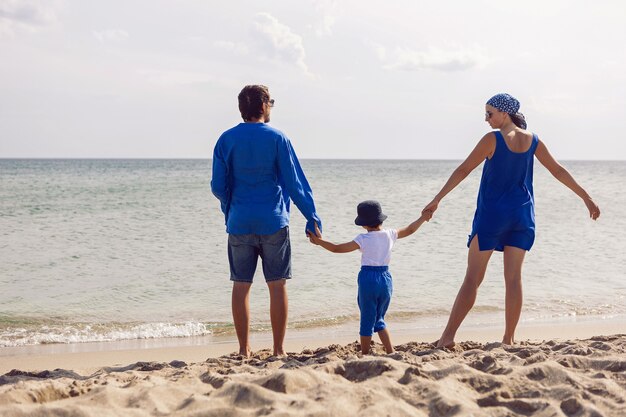 Familie van drie in blauwe kleren staat terug op een zandstrand aan zee in de zomer op vakantie