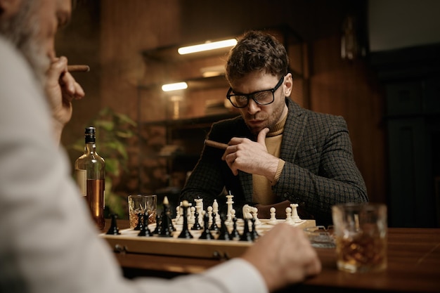 Familie van intelligente mensen die schaken, sigaren roken en whisky drinken. Senior en jongere mannen zitten aan tafel en kijken naar schaakbord. Slimme spellen