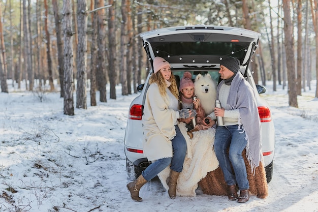 Familie van moeder, vader en dochter in warme jassen en een grappige witte Samojeed-hond zittend in de kofferbak van een auto in het winterbos. Gezinsgeluk, zorg voor de natuur