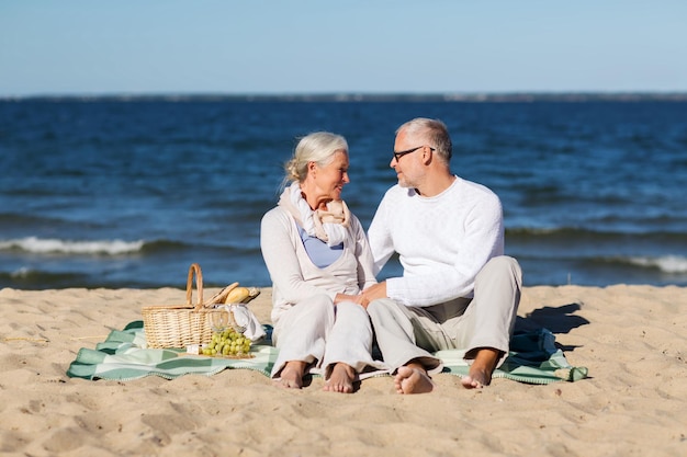 Фото Семья, возраст, праздники, досуг и концепция людей - счастливая пожилая пара с корзиной для пикника, сидящая на одеяле на летнем пляже