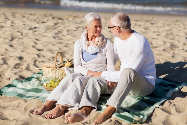 family, age, holidays, leisure and people concept - happy senior couple with picnic basket sitting on blanket on summer beach