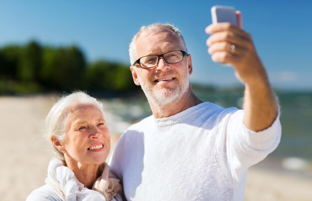 family, age, travel, technology and people concept - happy senior couple with smartphone taking selfie and hugging on summer beach