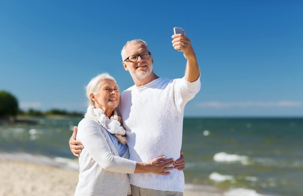 Family, age, travel, technology and people concept - happy senior couple with smartphone taking selfie and hugging on summer beach