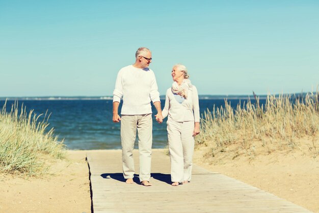 Фото Семья, возраст, путешествия, туризм и концепция людей - счастливая пожилая пара, взявшись за руки и прогуливаясь по летнему пляжу