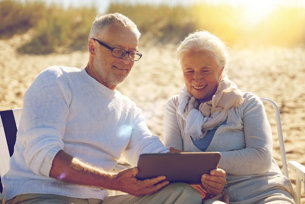 family, age, travel, tourism and people concept - happy senior couple with tablet pc computer resting in folding chairs on summer beach