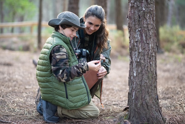 Family of amateur photographers in forest. Woman  and son in casual clothes with cameras kneeling near tree. Hobby, family, nature, photography concept