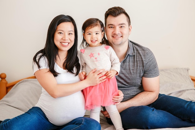 Family asian chinese pregnant woman and caucasian man with mixed race toddler girl sitting on bed