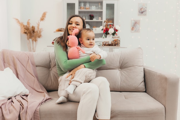 Family, baby and motherhood concept - happy smiling young mother with little baby on sofa at home. Mothers Day