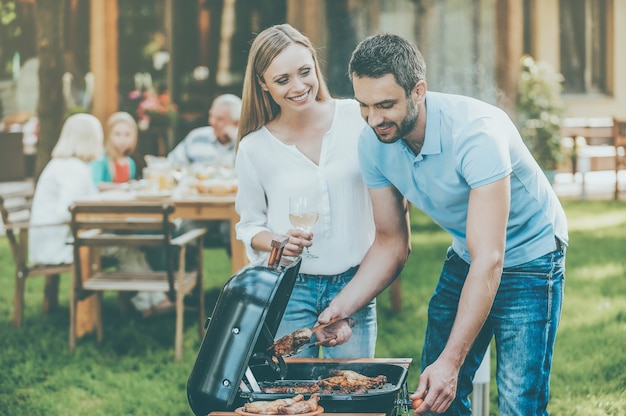 Family barbeque. happy young couple barbecuing meat on the\
grill while other members of family sitting at the dining table in\
the background