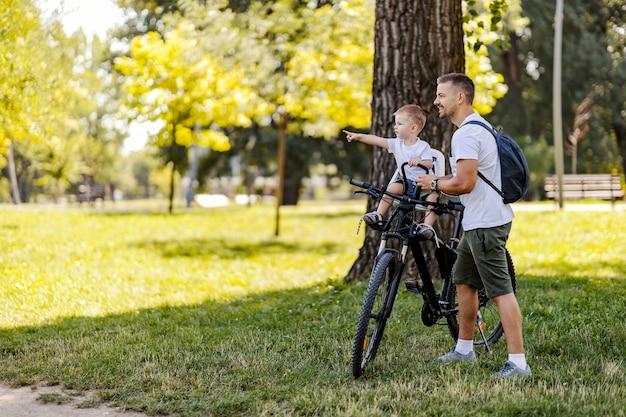 Family bike ride. Father and his son take a break from cycling in a green park on a sunny summer day. A toddler with a cap is sitting in a basket and pointing to something while man is standing