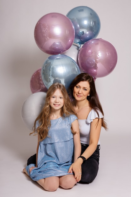 Family and birthday concept - mother and her daughter with pastel air balloons sitting over white background