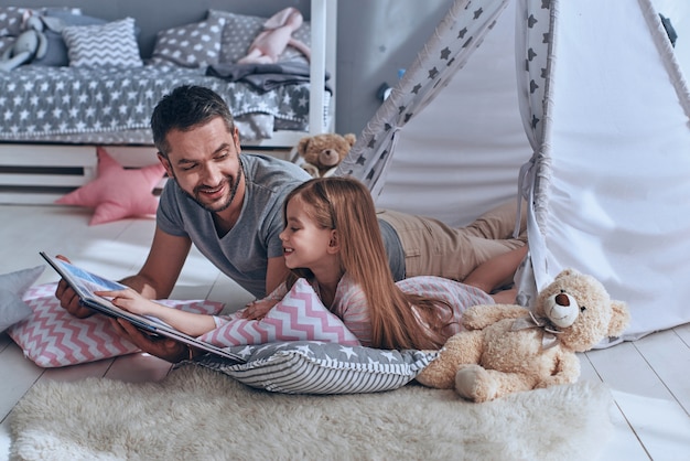 가족 결속. 침실 바닥에 누워 있는 동안 딸에게 책을 읽어주는 아버지